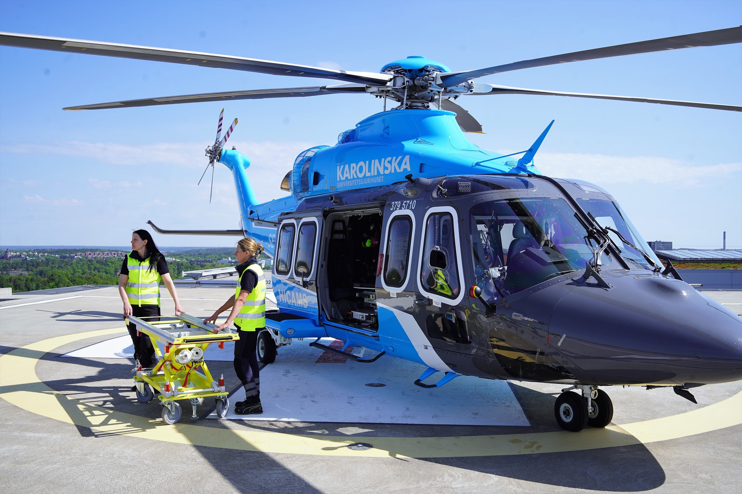 karolinska helikopter på helikopterplattan, två personer med patientbår står framför